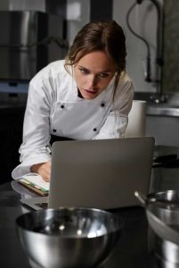 Köchin in der Küche mit Laptop-Gerät programm mitarbeitern bedienung rezeptverwaltung nutzung kassenbuch webshop